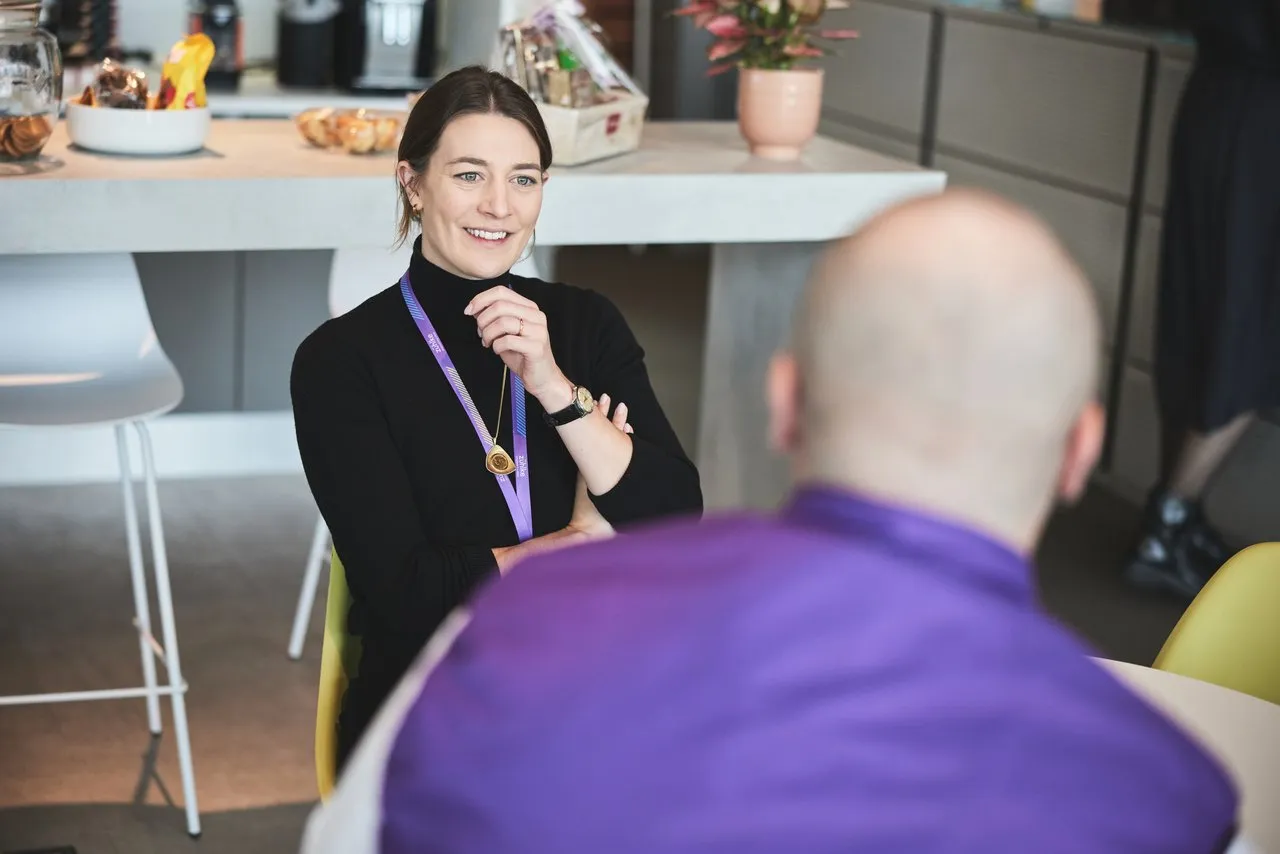 A woman sitting at the office kitchen table, smiling and talking to the colleague opposite of her