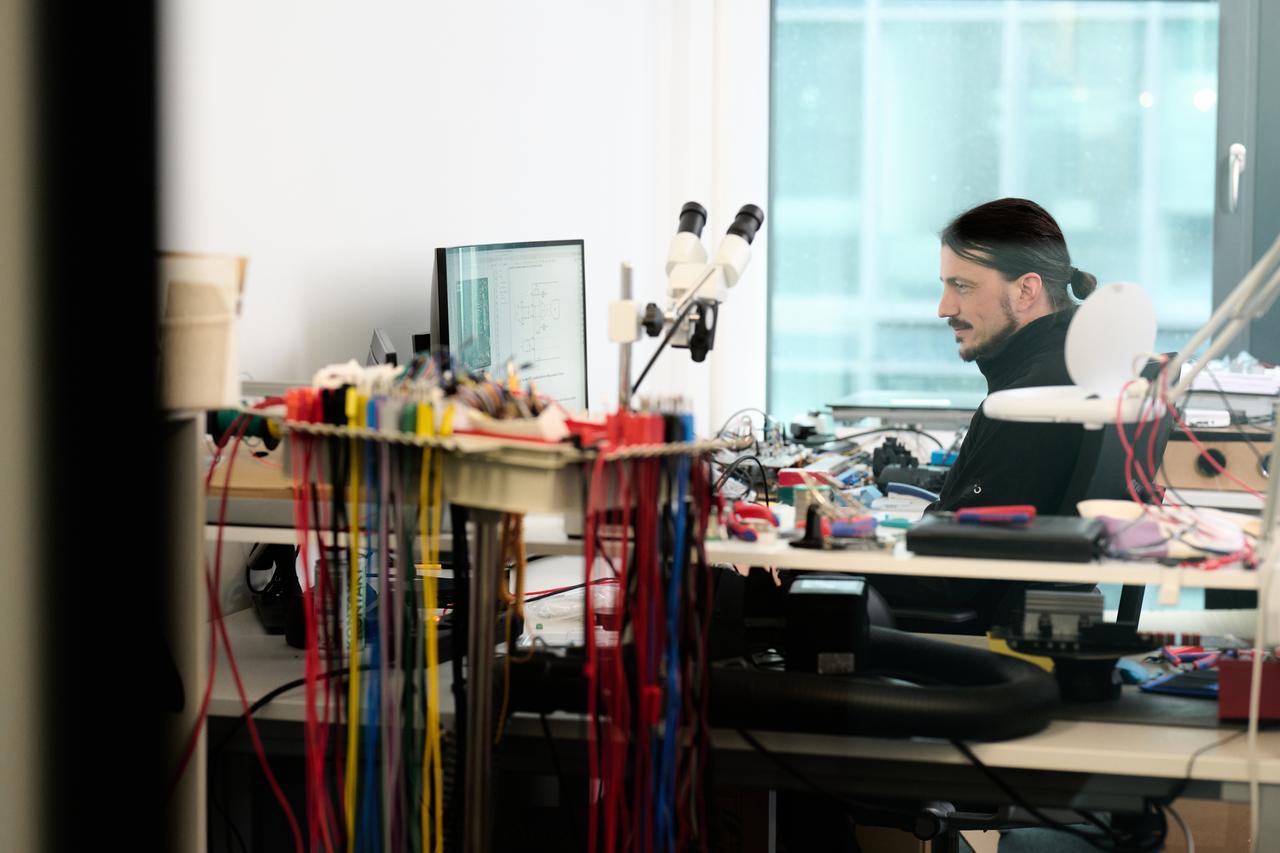 A man sitting in front of the computer screen at his desk surrounded by dzens of cables and connectors