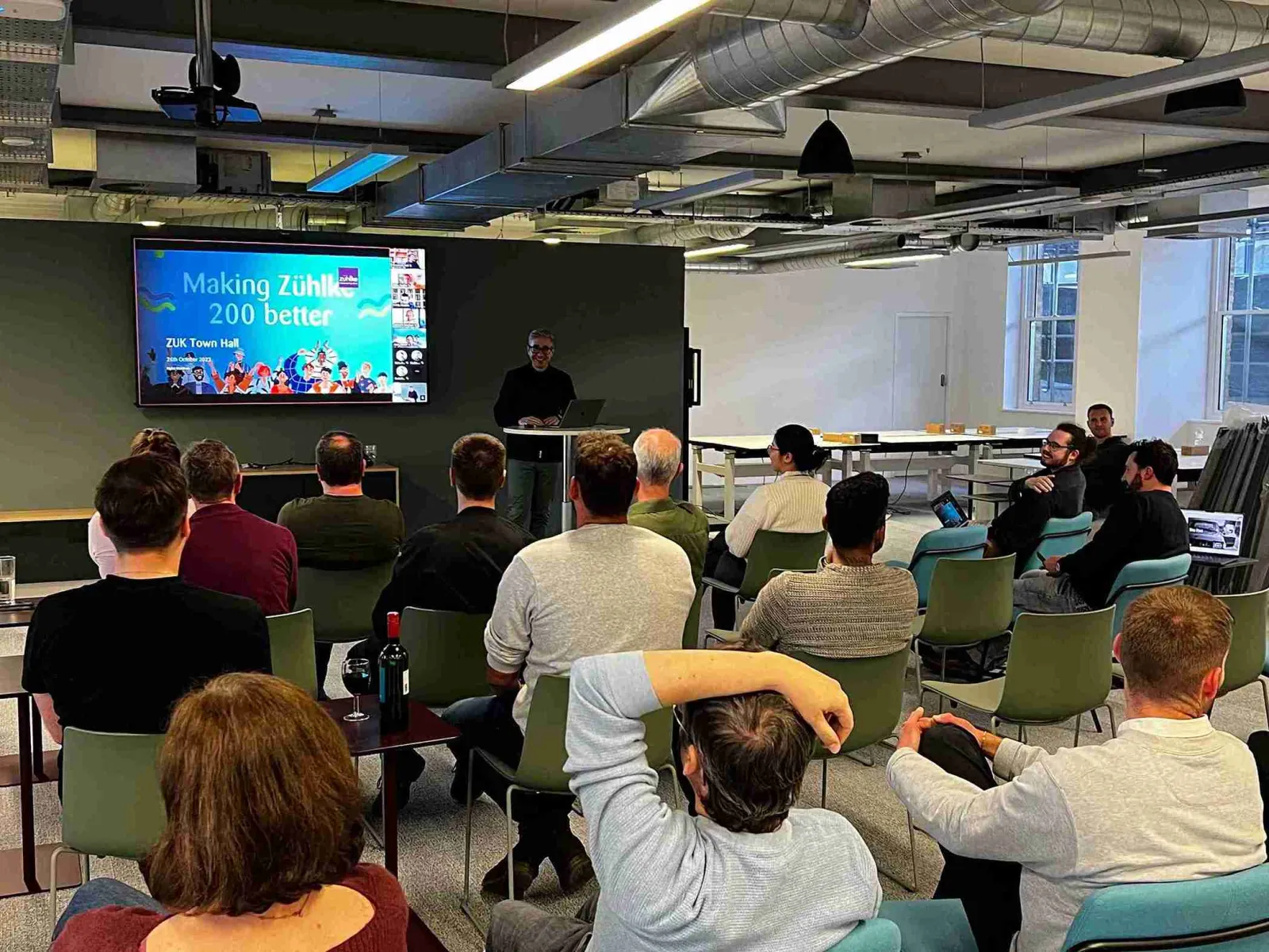 An office townhall event with people sitting in front of the digital screen and a presenter next to it