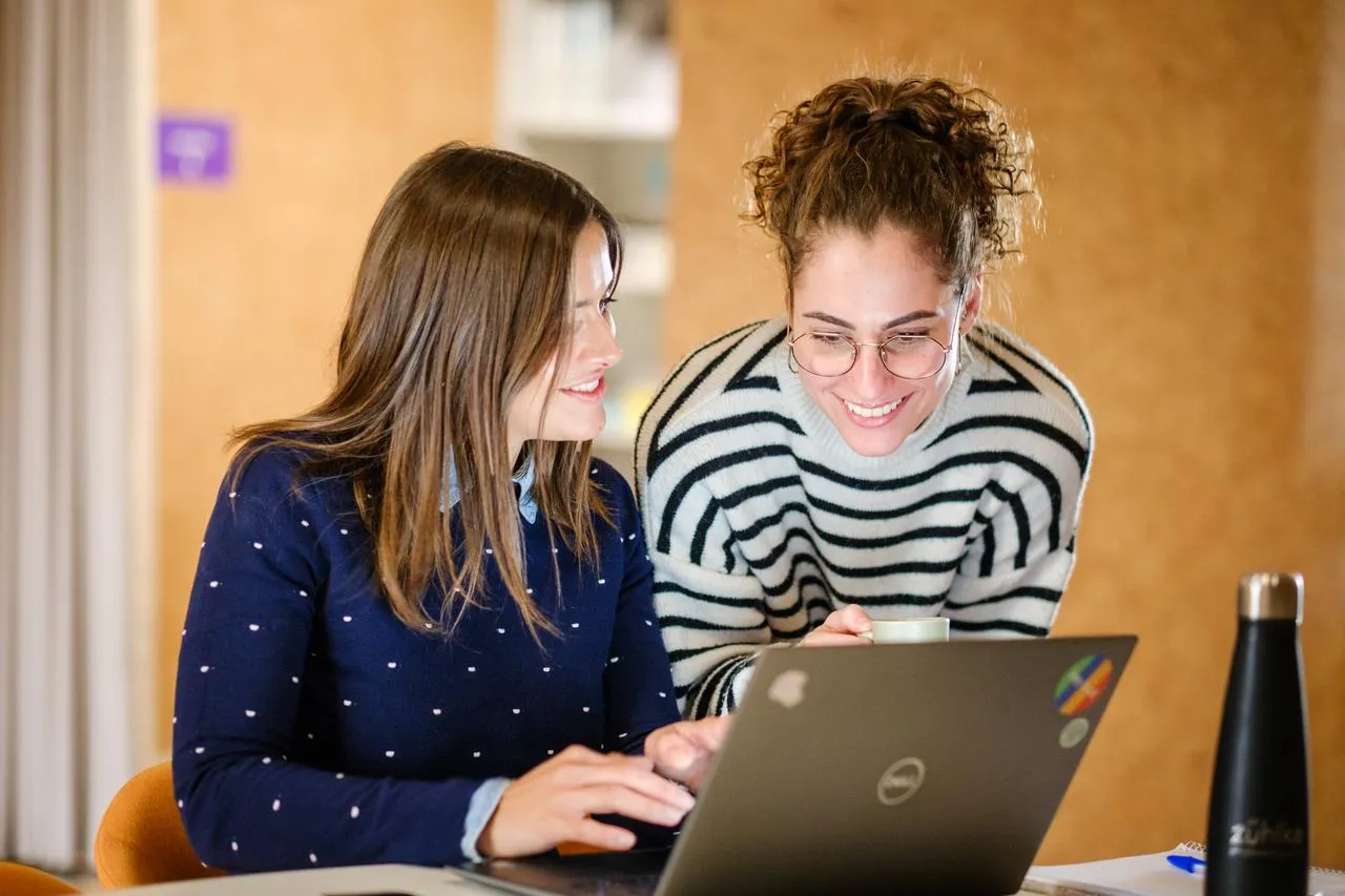 Two ladies at the office desk sitting and smiling in front of one laptop