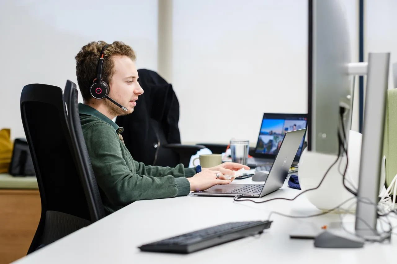 A software engineer in a headset sitting at the office desk in front of a laptop and monitor